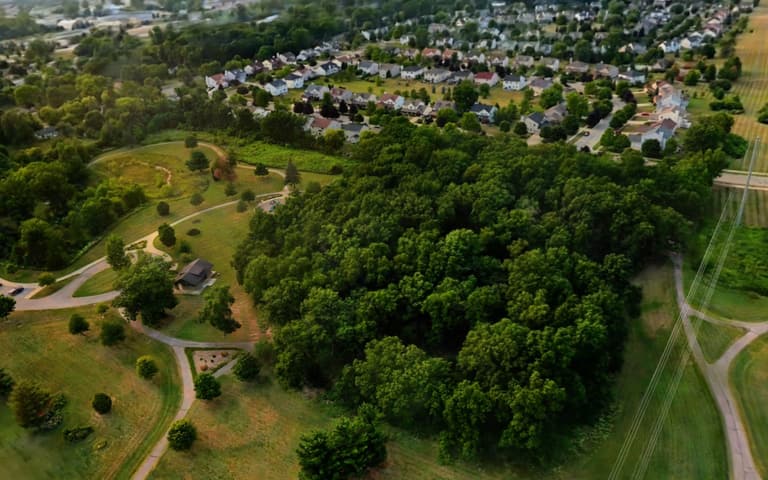 Live 3D model of a residential community from the air: streets of homes, a wooded hill, trails and a park shelter